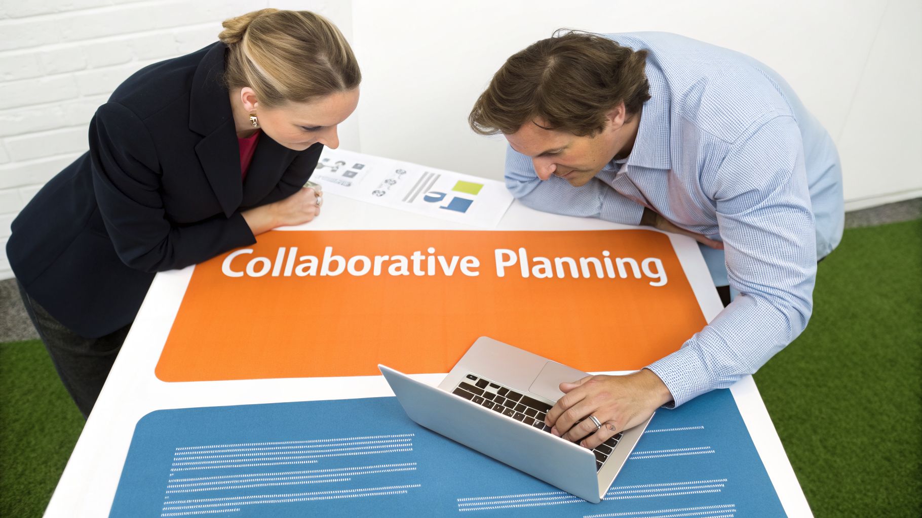 Two professionals engage in collaborative planning, working on a laptop and reviewing documents on a table.