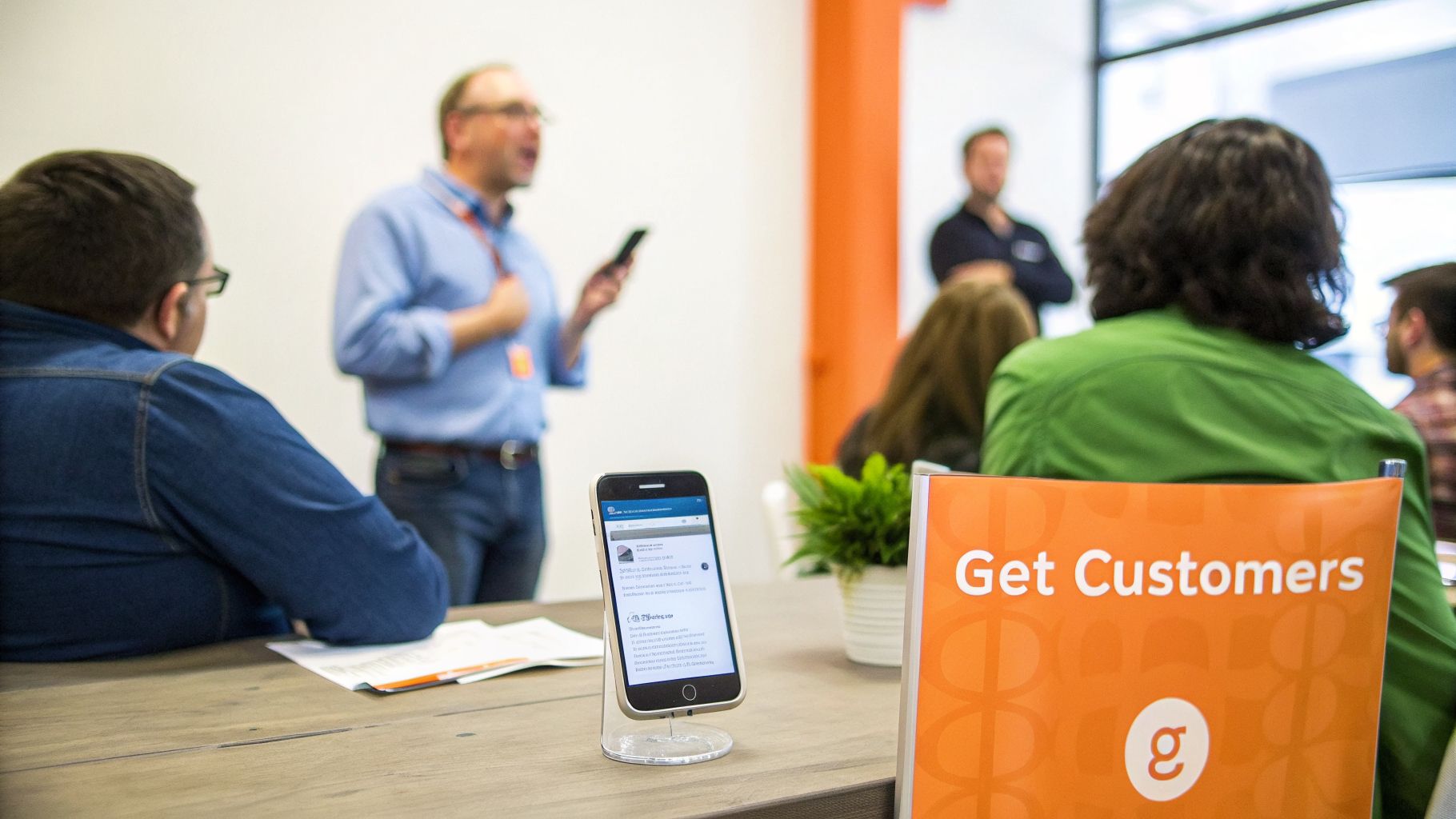 A speaker presents to attendees at a business workshop, with a smartphone displaying content on the table.