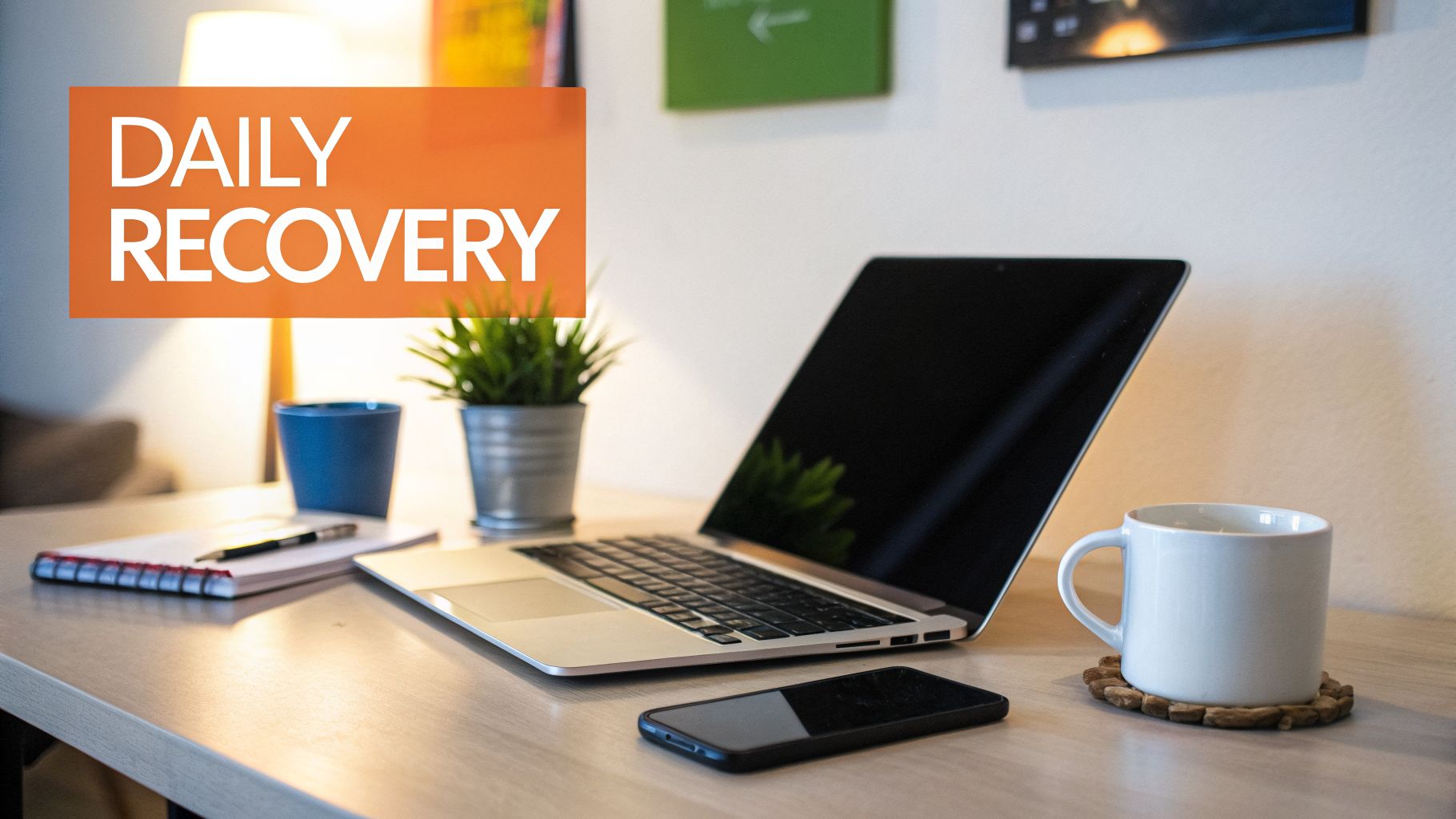 A brightly lit desk setup with a laptop, coffee mug, and an orange sign reading "DAILY RECOVERY".
