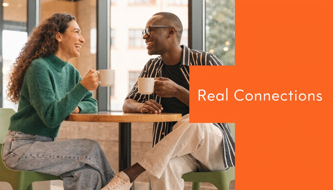 A smiling man and woman sitting at a table in a cafe, holding coffee mugs and talking.