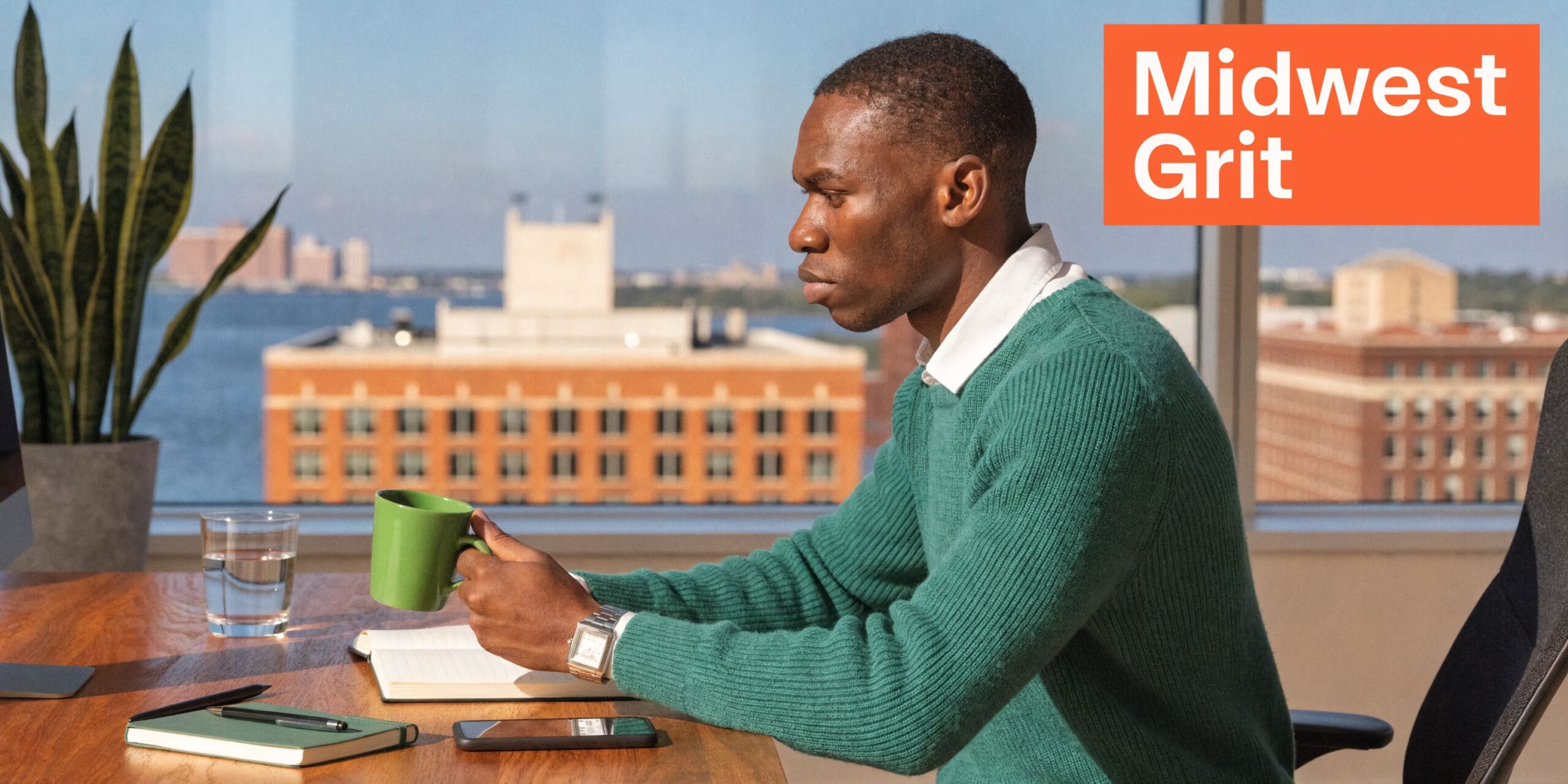 A professional man holding a green coffee mug while sitting at a sunny office desk overlooking a city.