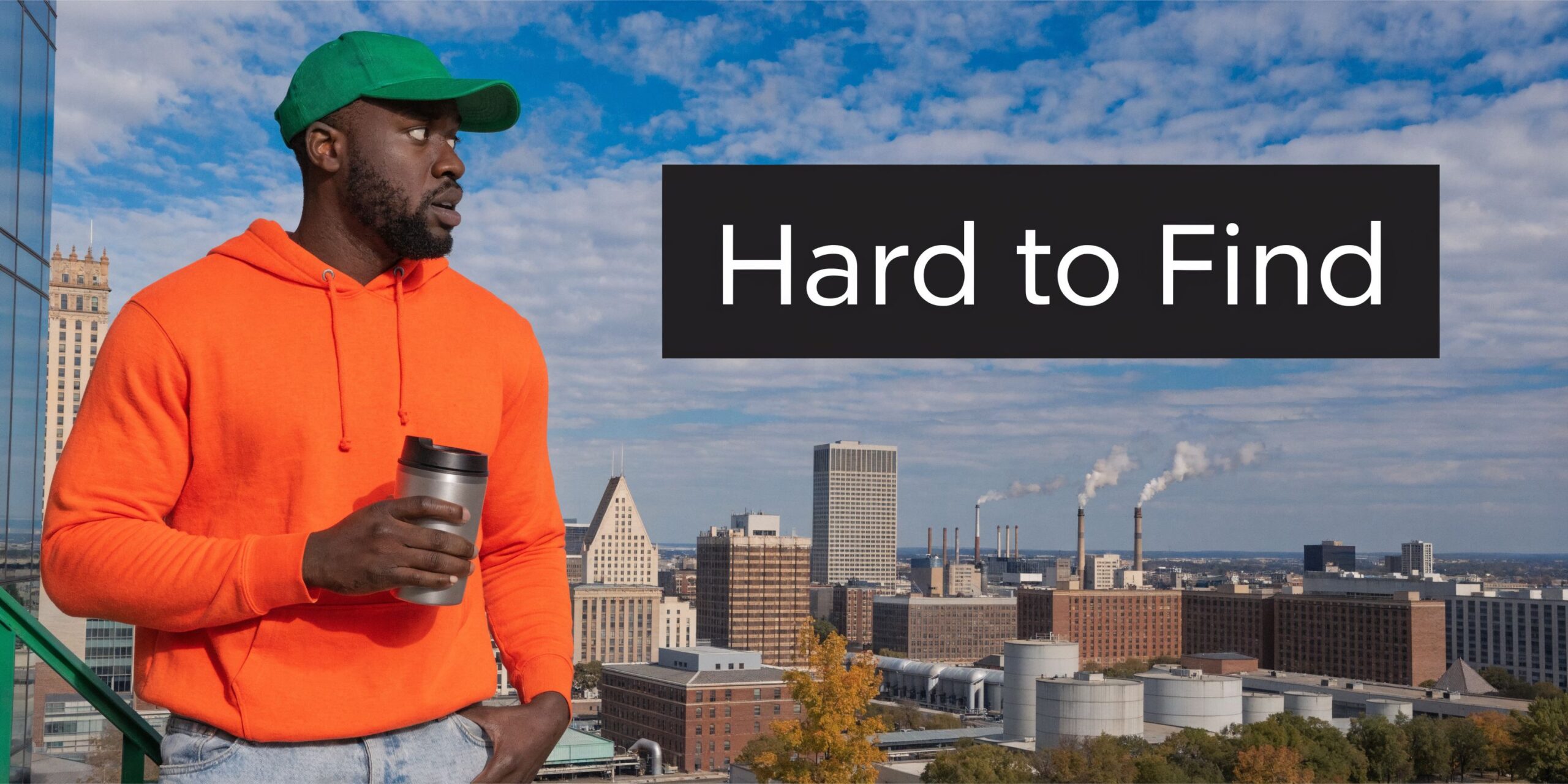 A man in an orange hoodie and green hat holding a cup overlooking industrial factory buildings.