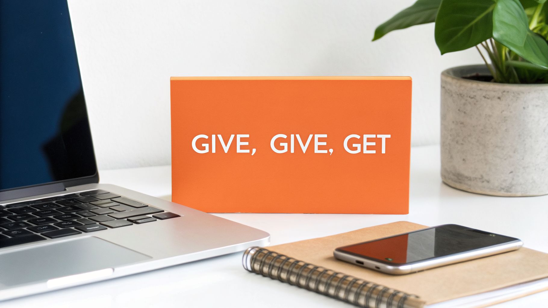 A modern desk setup featuring a laptop, smartphone, notebook, and an orange sign stating 'GIVE, GIVE, GET'.