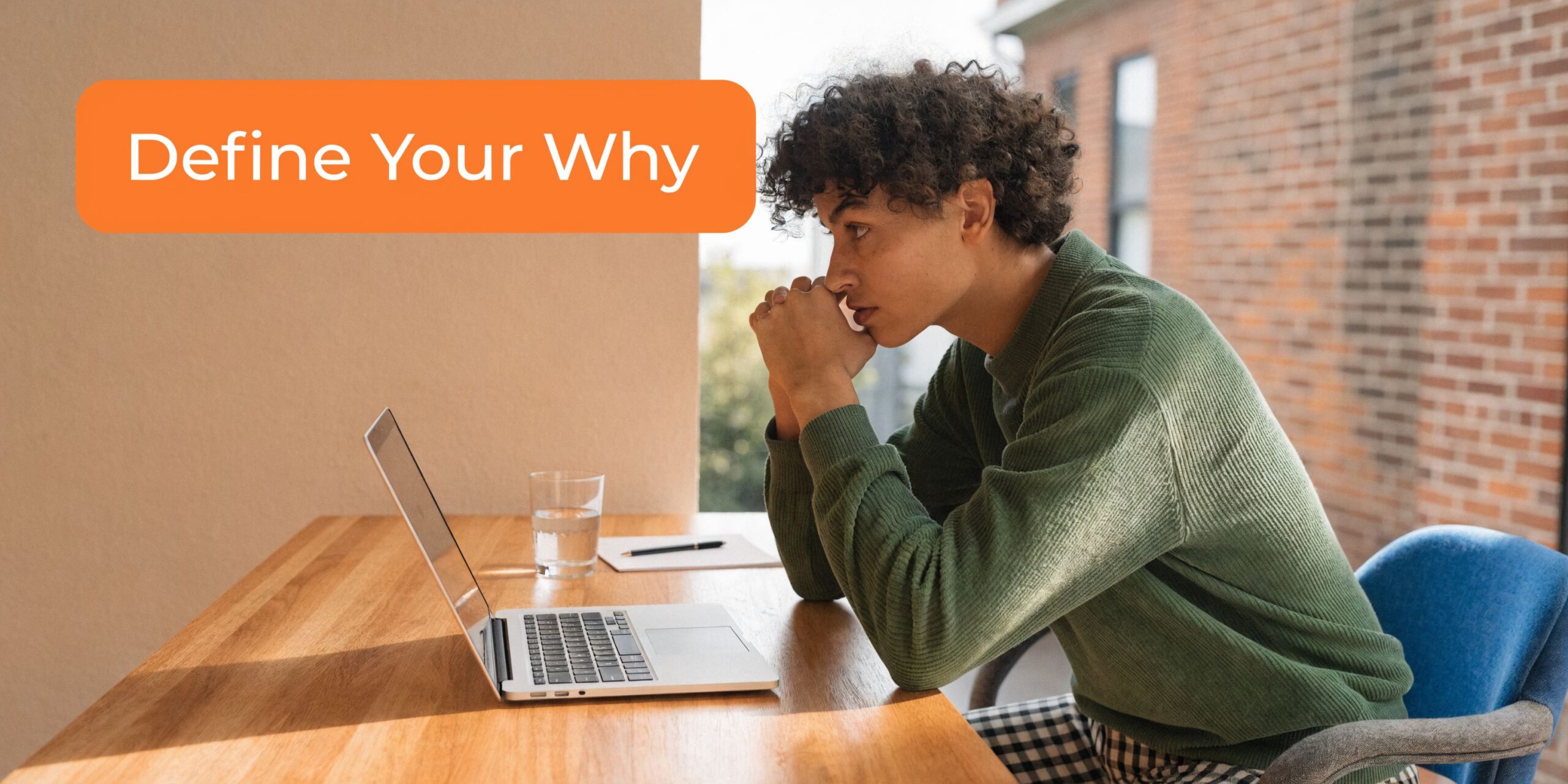 A young man with curly hair looking thoughtful at a laptop while sitting at a wooden desk.