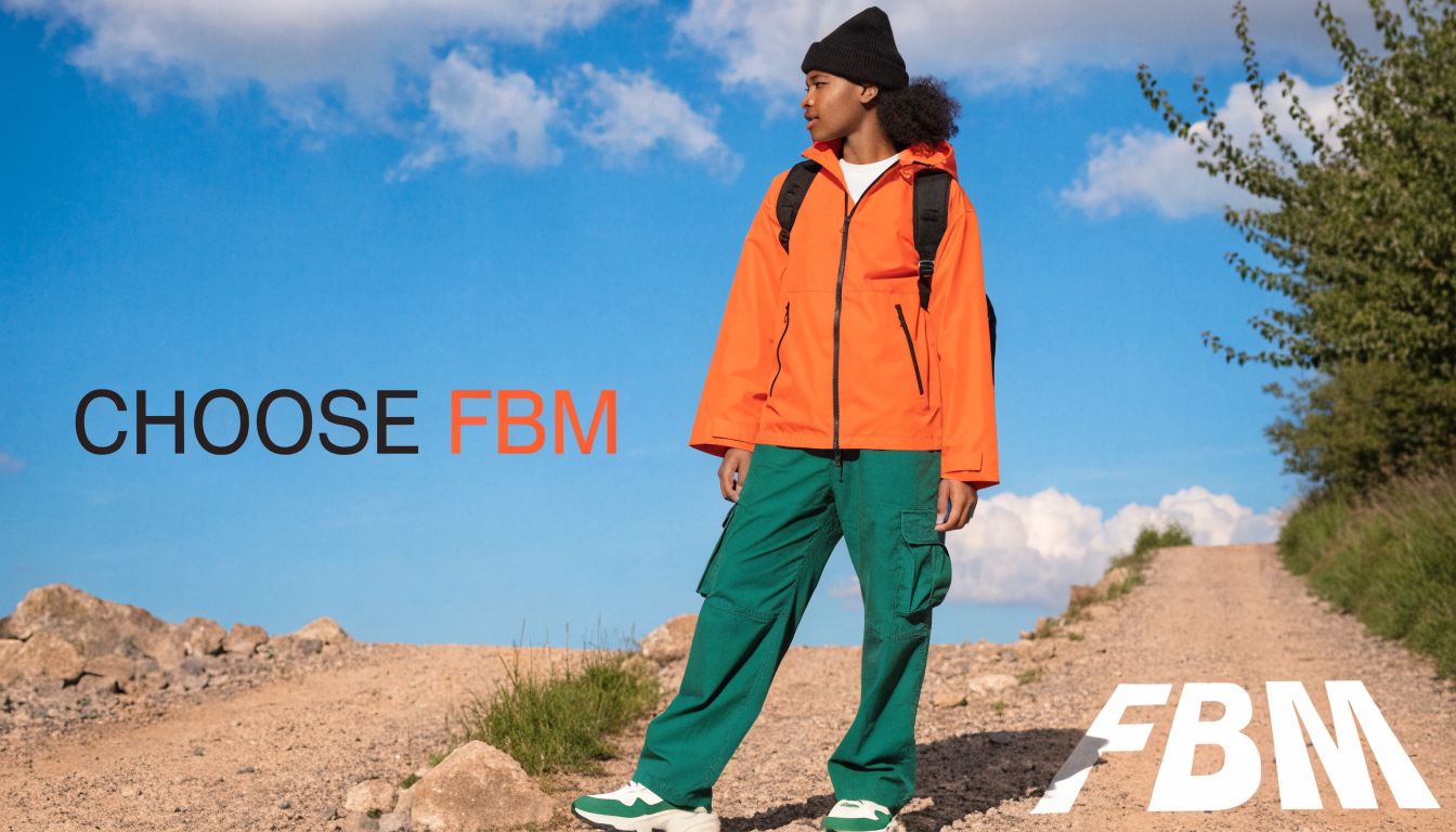 A person wearing a bright orange jacket and green cargo pants standing on a sunny dirt path.