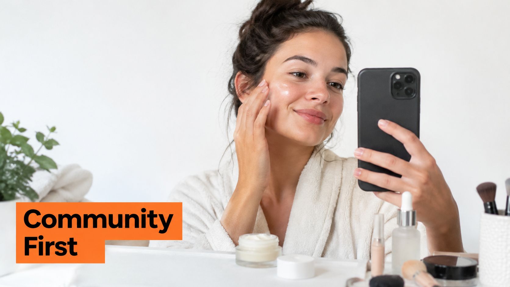 A woman in a white bathrobe applies facial cream while looking at her smartphone for a selfie.